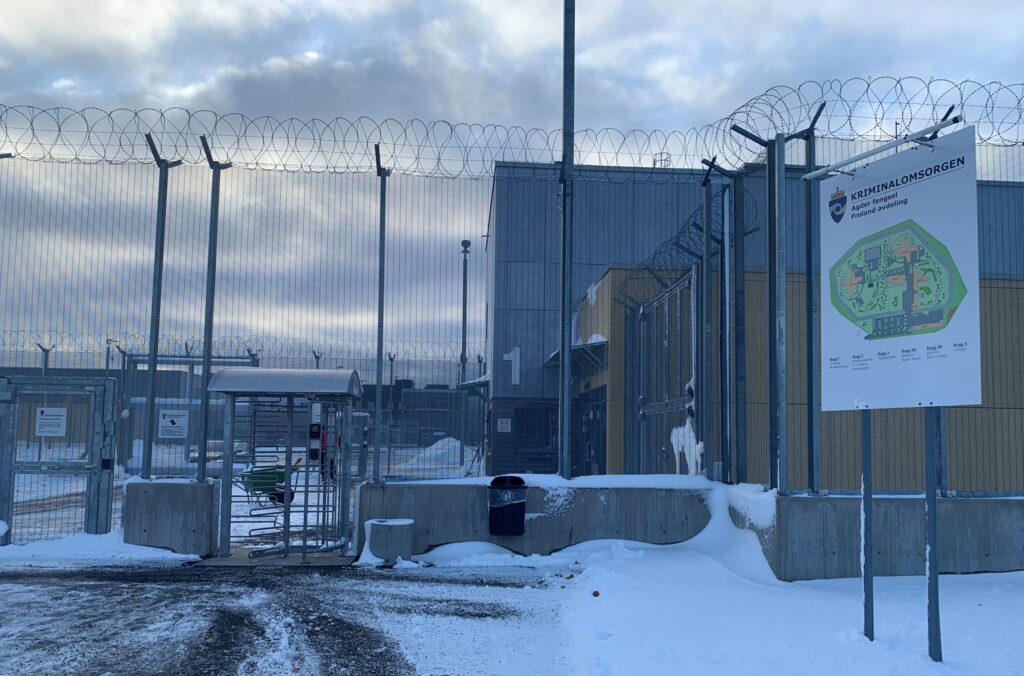 Bygniger bak høyt gjerde med piggtråd øverst. Skilt foran med oversiktskart over området og teksten 