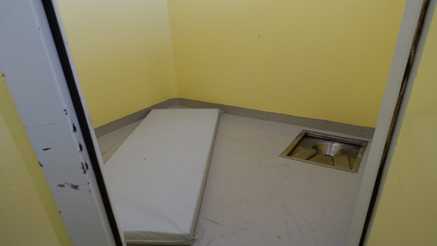 Interiør fengselscelle. Gul vegg, hull i golvet og madrass på golvet.