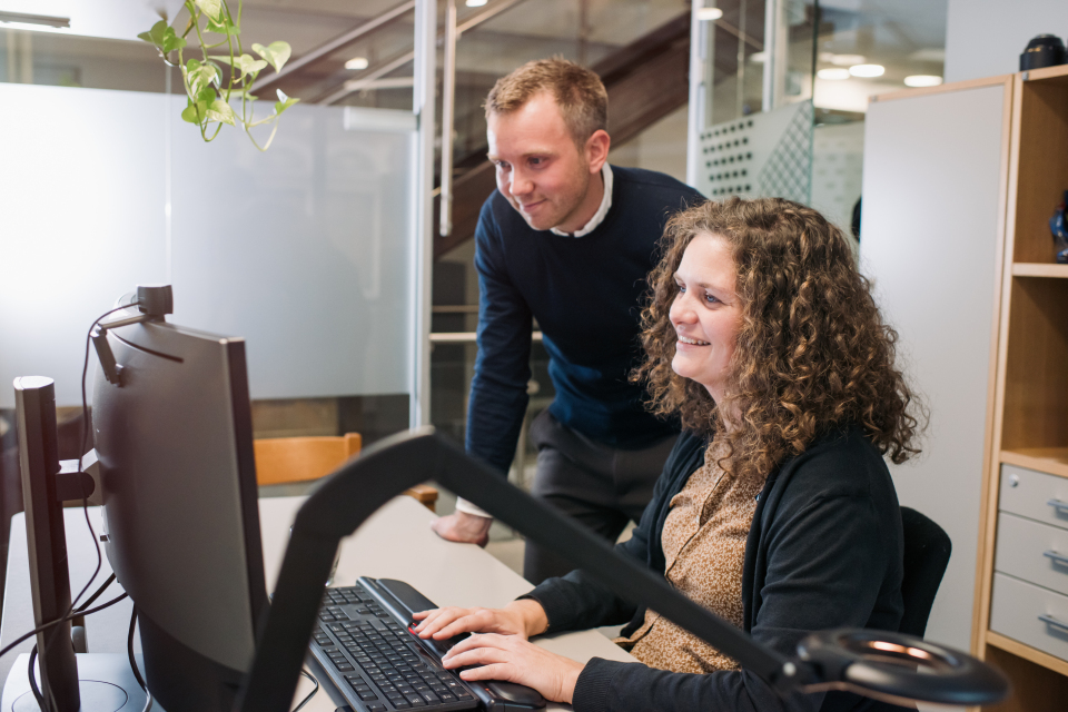 Arbeid på kontor. Kvinne sitter foran pc og mann står ved siden av og ser på skjermen.